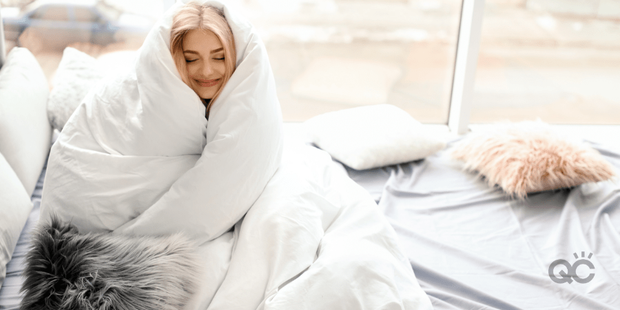 happy woman wrapped in blanket, sitting on bed, and smiling with content