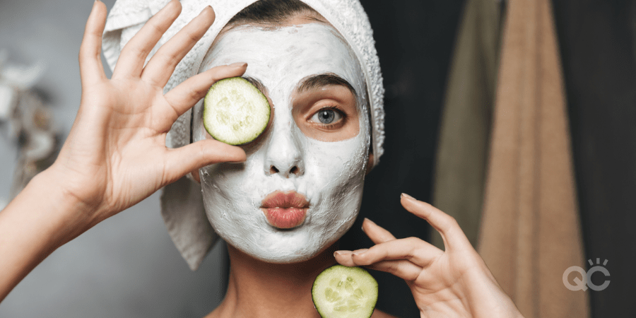 girl making kissy face, with hair up in towel, a face mask on, and cucumber slices in hand