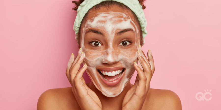 Smiling glad dark skinned young woman puts soap on face for cleansing, washes with cold water, wants to have healthy fresh skin., wears headband, looks positively at camera, stands naked indoor