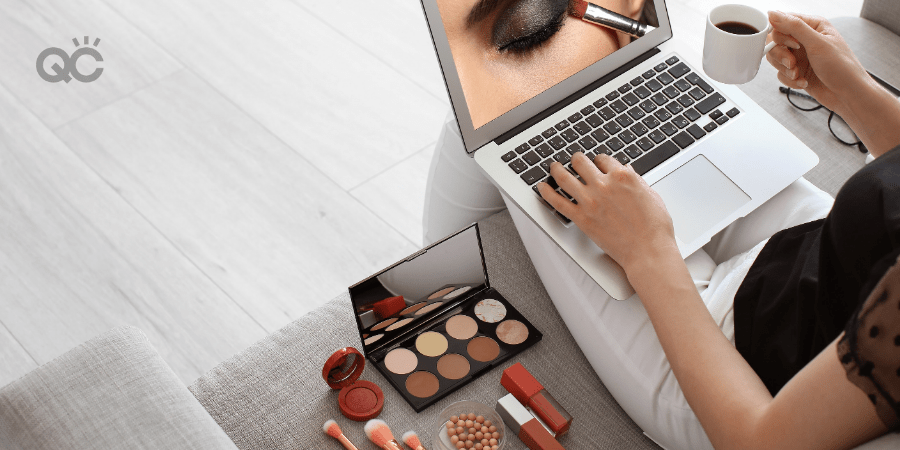 woman watching makeup lesson on laptop, sitting on couch with makeup products laid out next to her