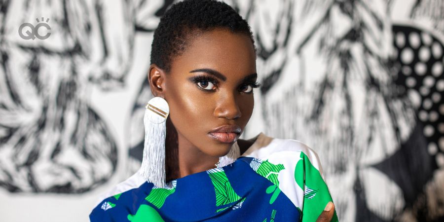 An elegant young black woman with short black hair & beautiful makeup and dangling earrings posing by herself indoors is wearing a pretty dress.