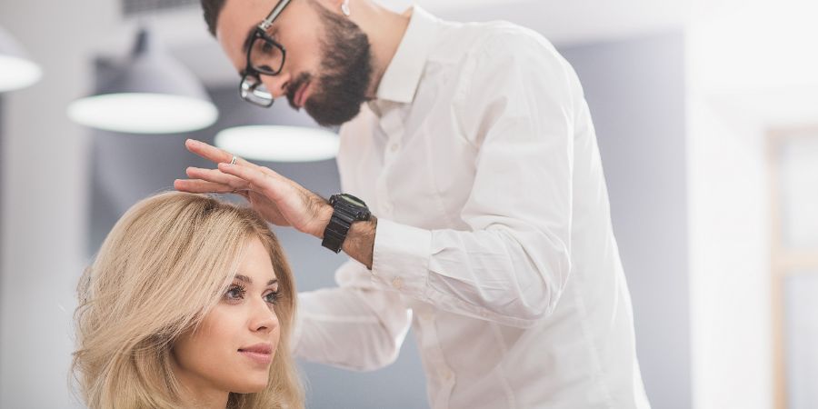 Male hair stylist working on blonde client in salon.