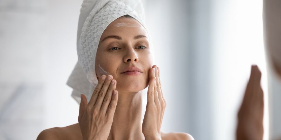 Closeup head shot pleasant beautiful woman applying moisturizing creme on face after shower. Smiling young pretty lady wrapped in towel smoothing perfecting skin, daily morning routine concept. Beauty habits article.