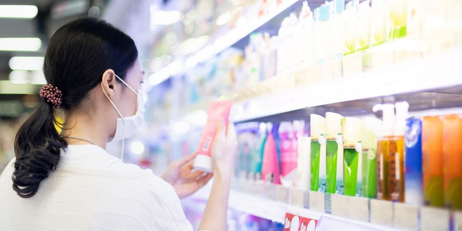 Asian woman wearing surgical mask and hand holding the bottle of facial foam for shopping in the aisle of the skincare department in supermarket, Due to the Covid-19 pandemic crisis. Skincare labels article.